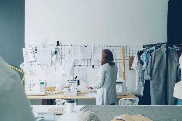 Designer trabalhando em um ateliê contemporâneo, cercado por máquinas de costura e projetos de roupas.