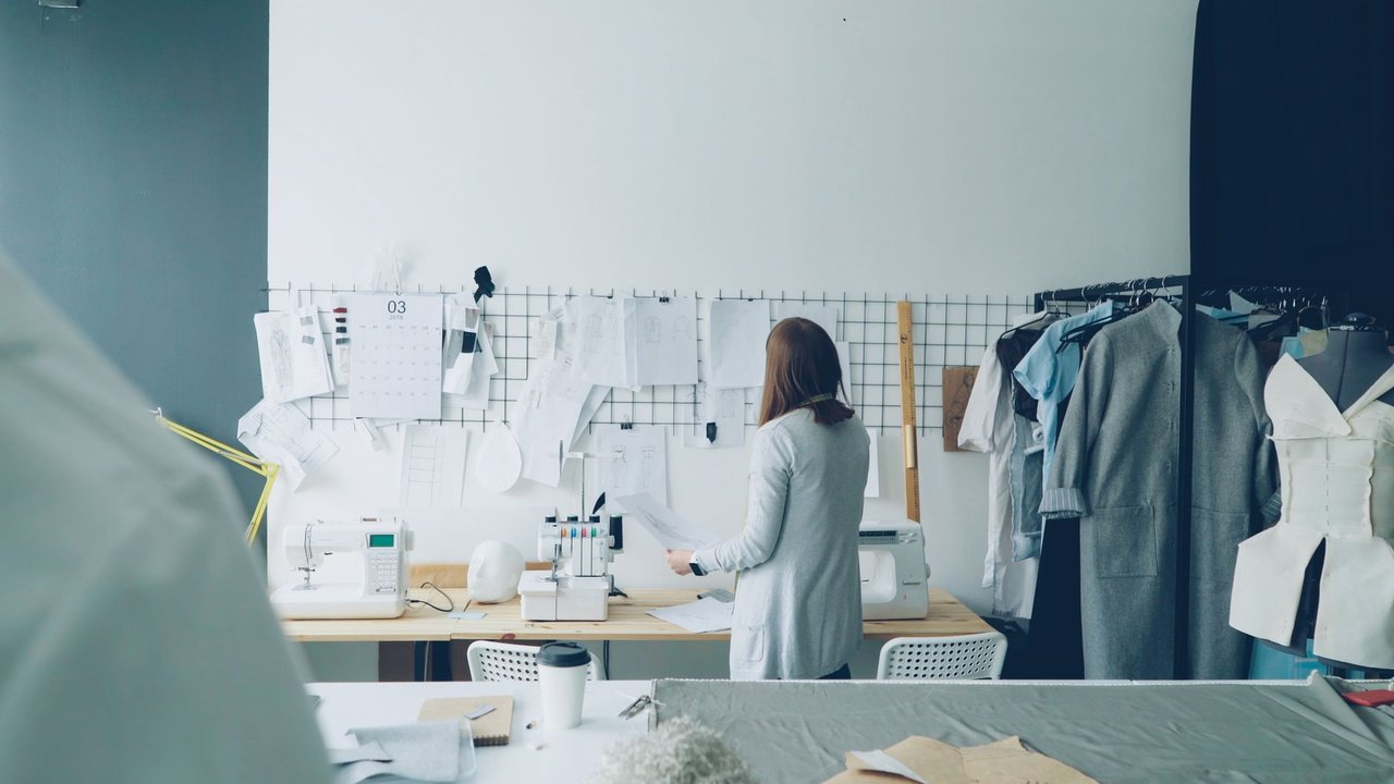 Designer trabalhando em um ateliê contemporâneo, cercado por máquinas de costura e projetos de roupas.