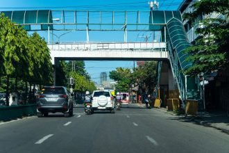 Passarela de pedestres sobre uma avenida urbana com veículos e circulação de pessoas.