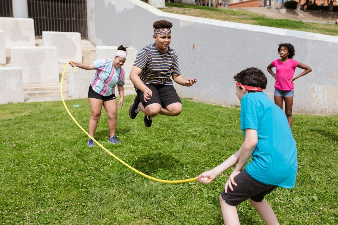 Grupo diverso de adolescentes brinca de pular corda ao ar livre, em um parque ensolarado.